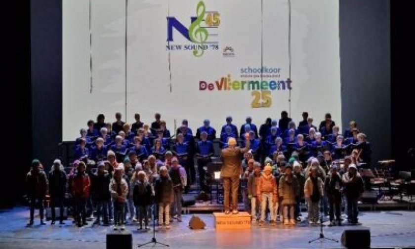 Op de afbeelding zie je het schoolkoor de Vliermeent en Zoetemeers gemengd koor 'New Sound '78'. Ze staan samen op een groot podium met in het midden dirigent Kees van Venetie.